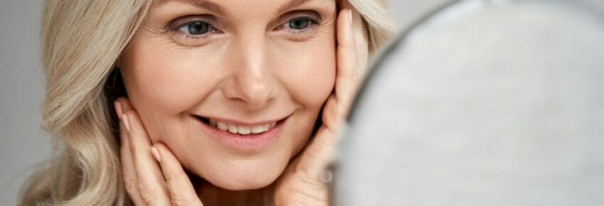 Happy 50s middle aged woman touching face skin looking in mirror.
