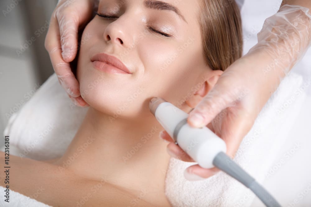 Close up of beautician hand making ultrasound facial procedure by special equipment. Her closed eyes with slight smile
